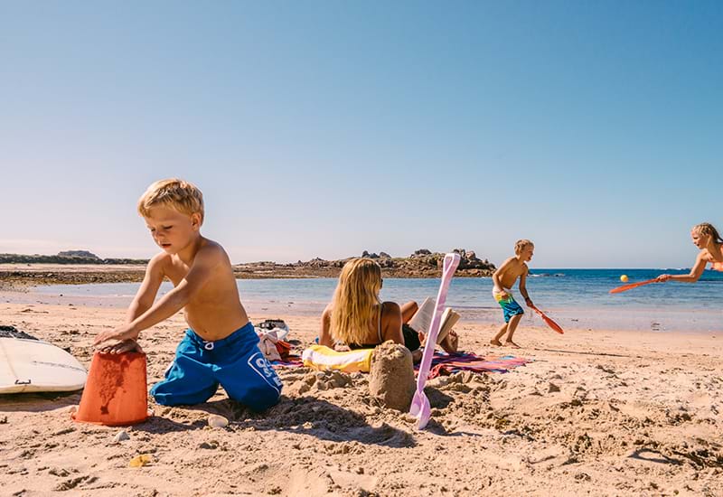 A child builds sandcastles on a beach with 2 children playing bat & ball in the background