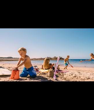 A child builds sandcastles on a beach with 2 children playing bat & ball in the background